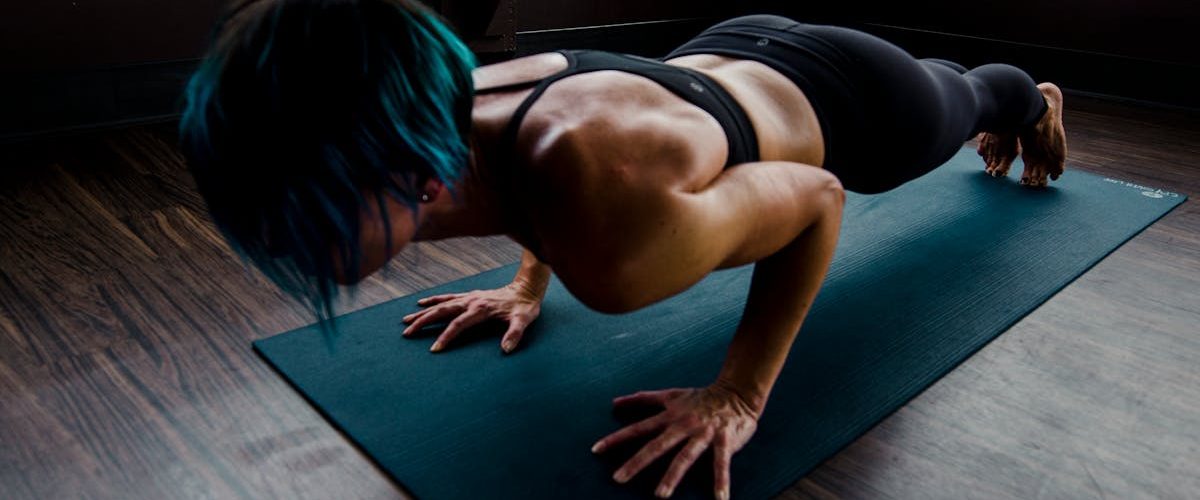 A fit woman with blue hair performs a push-up on a yoga mat indoors, showcasing strength and focus.